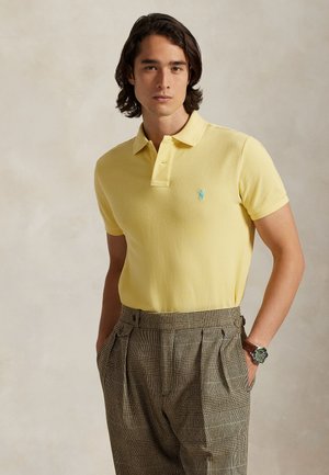 Άνδρας με μεσαίου μήκους μαλλιά που φοράει κίτρινη πόλο μπλούζα, καρό ψηλόμεσα παντελόνια και ένα ρολόι χειρός, με τα χέρια στις τσέπες, ουδέτερο φόντο.