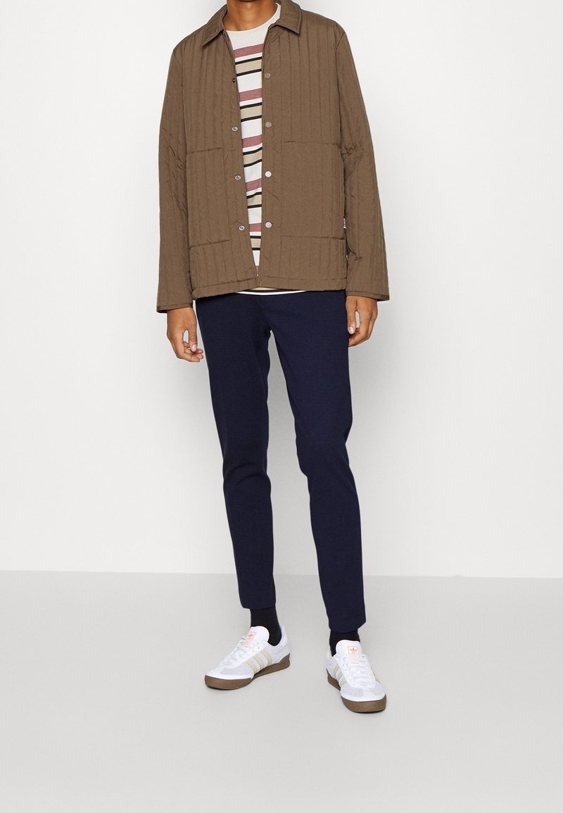 Mörkbrun vadderad jacka med tryckknappar, randig skjorta under, i kombination med marinblå byxor och vita sneakers med bruna accenter.