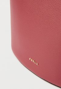 Geantă rotundă din piele burgundy texturată, cu logo auriu „Furla” în partea de jos, având o bază neagră netedă.