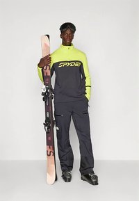 Abbigliamento da sci con maglia nera e verde neon con logo 'SPYDER', abbinata a pantaloni neri e sci rosa con attacchi neri in mano.