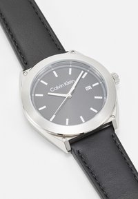 Reloj Calvin Klein con caja de acero inoxidable plateado, esfera gris, marcadores de hora plateados y correa de piel negra con detalle de costura.