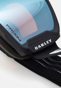 Zwarte en grijze skibril met een blauwgetinte lens, met het "Oakley" logo en de tekst "Prizm", bevestigd aan een zwarte band met witte accenten.