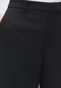 Gros plan sur la ceinture d’un pantalon formel noir avec passants pour ceinture et couture visible de la fermeture éclair frontale.