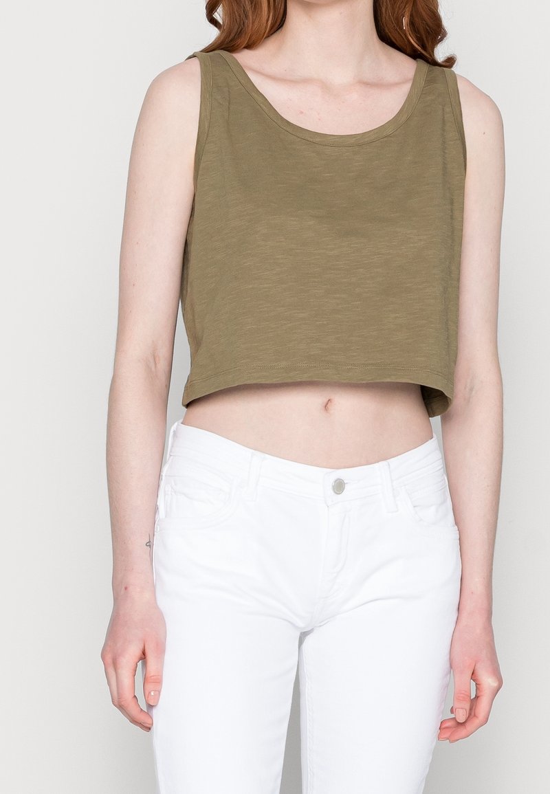 Personne portant un crop top sans manches vert olive et un pantalon blanc taille haute sur un fond clair uni.