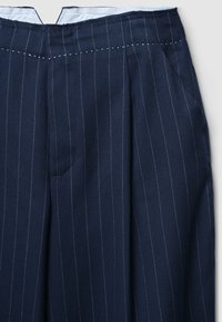 Pantalon bleu marine à fines rayures blanches verticales, devant plissé, poches latérales et doublure blanche à la ceinture.
