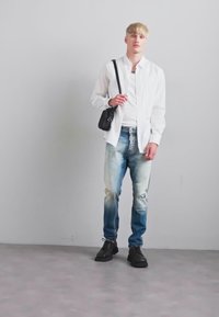 Młoda osoba z krótkimi blond włosami, nosząca białą koszulę, wyblakłe rozerwane dżinsy, czarne buty i trzymająca małą czarną torbę na ramię.