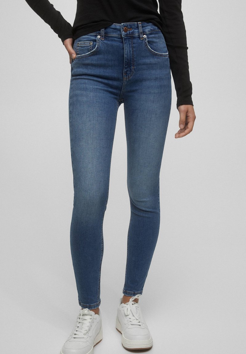 Persoon in mid-rise skinny blauwe jeans en witte sneakers tegen een effen lichtgrijze achtergrond.