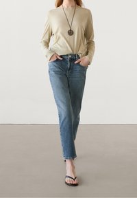 Vaalean beigen pitkähiomainen paita, joka on yhdistetty korkeavyötäröisiin vaaleansinisiiin farkkuihin. Mallilla on päällä mustat flip-flopit. Tausta on yksivärinen vaalean sävyinen.