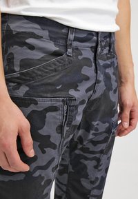 Närbild av en person som bär mörkgrå och svarta camouflageshorts och en vit skjorta, med händerna vilande längs sidorna.