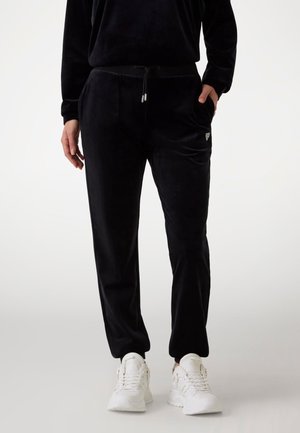 Persoon draagt zwarte velours joggerbroek met trekkoord in de taille en witte sneakers, staat met één hand in de zak.