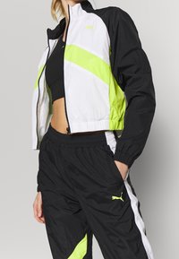 Woman wearing black sports bra, black and neon yellow Puma windbreaker jacket, and matching black track pants with white and neon yellow accents.