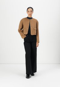 Bruine cropped jas met gouden knopen en twee voorkruikzakken, gedragen over een marineblauw topje. Gecombineerd met zwarte wijde broek en zwarte schoenen.