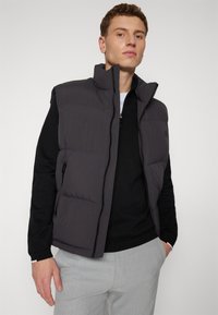 Puffer vest in dark gray fabric, featuring a high collar, zip closure, and side pockets, layered over a black top. Lightweight and quilted texture.