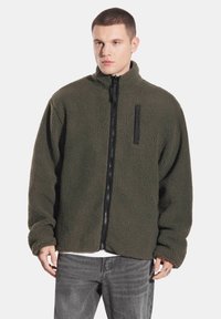 Błyszcząca oliwkowo-zielona kurtka polarowa z wysokim kołnierzem, zamkiem błyskawicznym z przodu i teksturowaną powierzchnią. Posiada czarną kieszeń na piersi zapinaną na zamek.