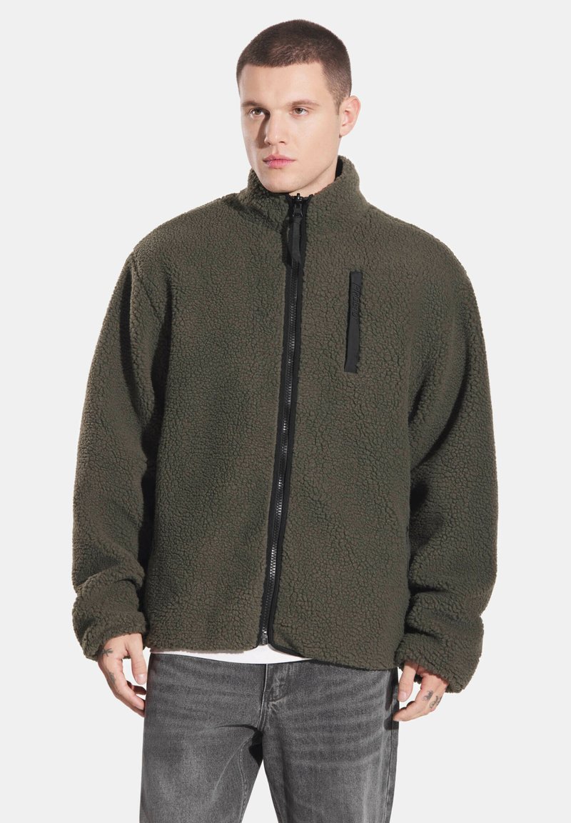 Błyszcząca oliwkowo-zielona kurtka polarowa z wysokim kołnierzem, zamkiem błyskawicznym z przodu i teksturowaną powierzchnią. Posiada czarną kieszeń na piersi zapinaną na zamek.