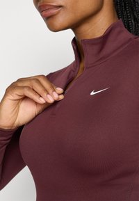 Burgunderfarget treningsgenser med kvart glidelås laget av glatt stoff. Har høy krage og en liten hvit Nike-logo på brystet.