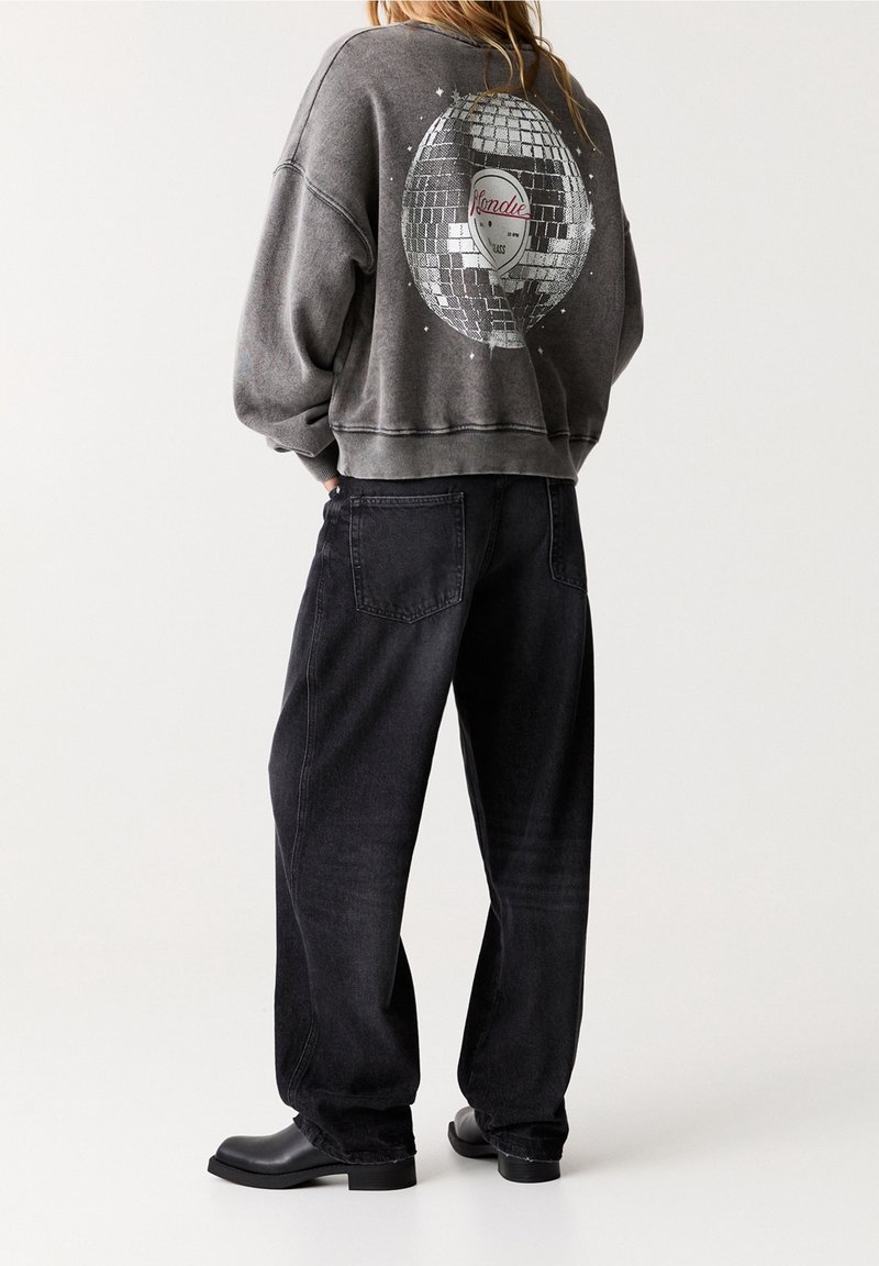 Femme debout, les mains dans les poches, portant un sweat gris avec un grand motif de boule à facettes et un jean noir à jambes larges.