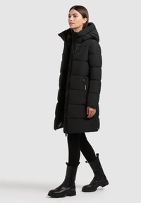 Black puffer coat with a hood, featuring a quilted design, zippered front pockets, and a mid-length cut. Paired with black boots.