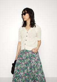 Cardigã de malha em marfim com mangas curtas e botões dourados, combinado com uma saia floral multicolorida com padrões em verde, rosa e branco.