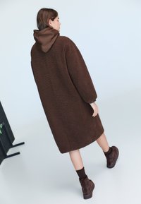 Ruskea, teksturoitu takki hupulla, jossa pitkät hihat ja rento istuvuus. Yhdistetty tummanruskeisiin nilkkureihin ja resorisiin sukkiin.