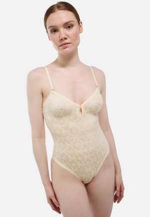 Femme portant un body en dentelle beige à motifs floraux avec de fines bretelles, debout devant un fond uni gris clair.