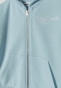 Svijetloplava dukserica od mekanog materijala, sa zatvaranjem na zatvarač, prednjim džepom i vezom "Paris Tennis Club".