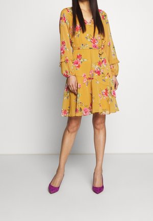 Robe fleurie en jaune moutarde avec des manches longues, des poignets volantés, une ceinture à nouer à la taille et une jupe fluide. Associée à des talons violets.