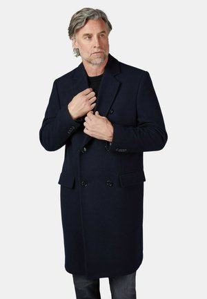 Middelbare man met grijs haar die een donkerblauwe dubbelrijige jas draagt over zwarte kleding, terwijl hij de kraag van de jas verstelt.