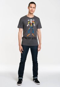 Graues Baumwoll-T-Shirt mit einem Cartoon-Charakter, der einen Cowboyhut trägt und leuchtende Farbakzente hat, kombiniert mit dunklen Jeans und schwarzen Turnschuhen.