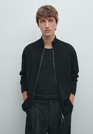 Svart stickad zip-up cardigan med hög krage, ribbade ärmslut, bärs över en svart T-shirt, tillsammans med skräddarsydda svarta byxor.