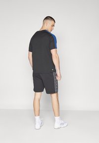 Črna kratka majica s kratkimi rokavi z modrimi detajli, v kombinaciji s črnimi kratkimi hlačami z belimi stranskimi črtami, obuta v bele športne čevlje.