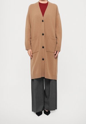 Femme portant un long cardigan camel avec poches avant, boutons noirs, un top rouge, un pantalon large gris et des chaussures noires pointues.