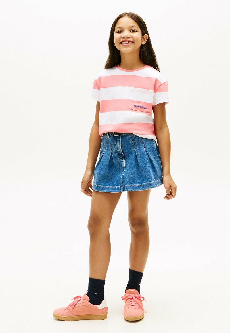 Fille souriante portant un t-shirt rayé rose et blanc, une jupe en jean bleue, des chaussettes noires et des baskets roses, debout sur un fond blanc uni.