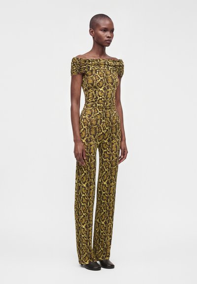 Femme portant une combinaison épaules dénudées jaune et marron à imprimé serpent, pantalon évasé et chaussures noires, debout devant un fond uni.