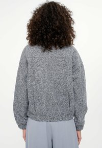 Osoba z kręconymi włosami, ubrana w teksturowany szary sweter z długim rękawem oraz luźne jasnoszare spodnie, odwrócona tyłem na białym tle.