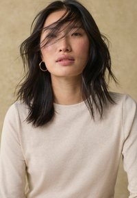 Maglia a maniche lunghe beige chiaro con finitura testurizzata. Il modello ha capelli scuri e ondulati con luci sottili. Orecchini a cerchio in oro completano il look.