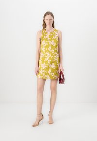 Rochie florală de culoare galbenă cu flori roz, design fără mâneci, croi drept, textură fină. Modelul ține o geantă roșie, purtând pantofi cu toc bej.
