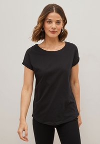 Femme aux cheveux bruns mi-longs, portant des boucles d'oreilles argentées et un t-shirt noir uni à manches courtes sur un fond clair.