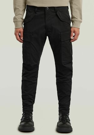 Mann trägt schwarze Cargohosen mit mehreren Taschen, beiges Langarmshirt und schwarze Schnürstiefel, steht vor einem einfarbigen Hintergrund.