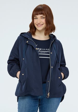 Junge Frau mit kurzen roten Haaren, die eine marineblaue Kapuzenjacke und Jeans trägt, lächelnd mit den Händen in den Jackentaschen.