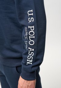 Mörkblå sweatshirt med vittryckt text på vänster ärm som säger "U.S. POLO ASSN. SINCE 1890." Mjuk bomullsmaterial, avslappnad passform.