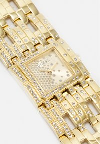 Gouden horloge met een vierkante wijzerplaat, bezaaid met rhinestones. Heeft een gestructureerde wijzerplaat en een schakels design armband.