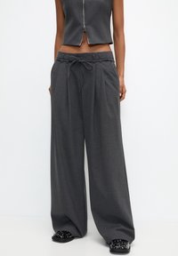 Grijze wijde broek met een koord in de taille, een geplooid voorpaneel en zachte stof. Draagt bij zwarte schoenen met studs.