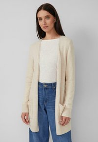 Donna che indossa un cardigan beige aperto con tasche anteriori sopra una maglietta bianca e jeans a vita alta blu, su uno sfondo grigio semplice.