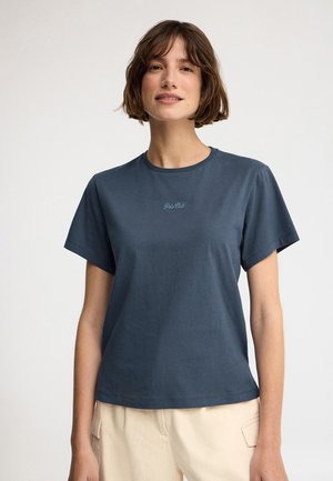 Joven con cabello corto y castaño, vistiendo una camiseta azul de Polo Club y pantalones beige claros, posando frente a un fondo claro y liso.