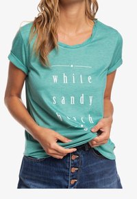 T-shirt in cotone turchese con maniche arrotolate, con la scritta bianca "spiaggia di sabbia bianca". Morbido al tatto, vestibilità rilassata, abbinato a pantaloni corti in denim.