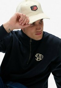 Mørkeblå zip-up sweatshirt med beige broderet logo på brystet, parret med en cremefarvet baseballkasket med et design af en sort og rød badge.