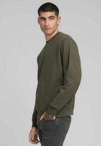 Felpa verde oliva con maniche lunghe, polsini a costine, scollo rotondo e vestibilità comoda. Presenta una texture liscia e un design minimalista.