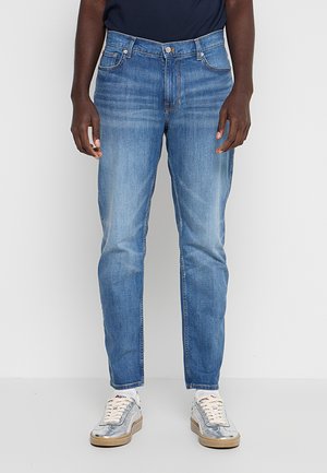 Uomo con jeans slim fit azzurri chiari, calzini bianchi e sneakers argento in piedi davanti a uno sfondo grigio semplice.