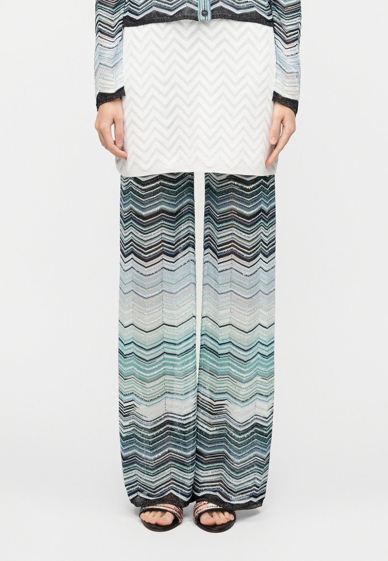 Večbarvne hlače s šivanim vzorcem v odtenkih modre, zelene in črne, kombinirane z belo zgornjo krinko z zigzagnim teksturo. Vidni so ravni sandali.